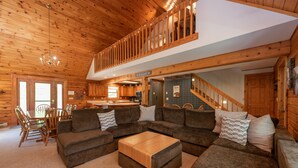 A spacious living room with a large sectional sofa, a wooden coffee table, and a dining area with a table and chairs. A staircase leads to an upper level with a balcony. - Enjoy Adventure & Relaxation w/ Hot Tub & Fire Pit (McHenry)