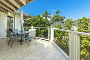 Un balcon avec une table et des chaises pour prendre les repas, un parasol blanc et une vue sur des arbres tropicaux.