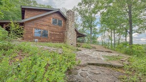 A rustic cabin with a stone chimney, surrounded by lush greenery and a stone pathway leading to the entrance. - Log Cabin right next to entrance of the Great Smokey Mountains National Park! (Waynesville)