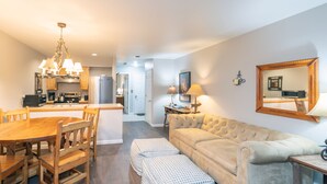 A modern living room with a beige sofa, wooden dining table, and matching chairs. There is a kitchen area with a refrigerator, stove, and microwave. A large mirror hangs on the wall, and a chandelier provides lighting. - Cozy Condo steps to Coonskin lift (Telluride)