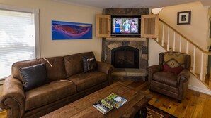 Ein Wohnzimmer mit einem braunen Ledersofa, einem Couchtisch mit Zeitschriften, einem Steinkamin und einem Fernseher auf einem Holzständer.