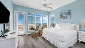 A bedroom with a large bed, a ceiling fan, and a view of the beach through the windows.