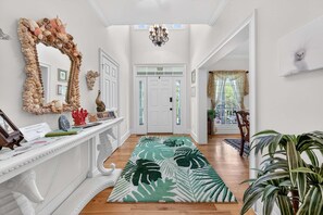 A hallway with a white console table, a mirror with a shell frame, a tropical-themed rug, and a chandelier.