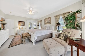 A bedroom with a bed, bedside tables, a chair, a desk, a floor lamp, a ceiling fan, a window with curtains, and a plant.