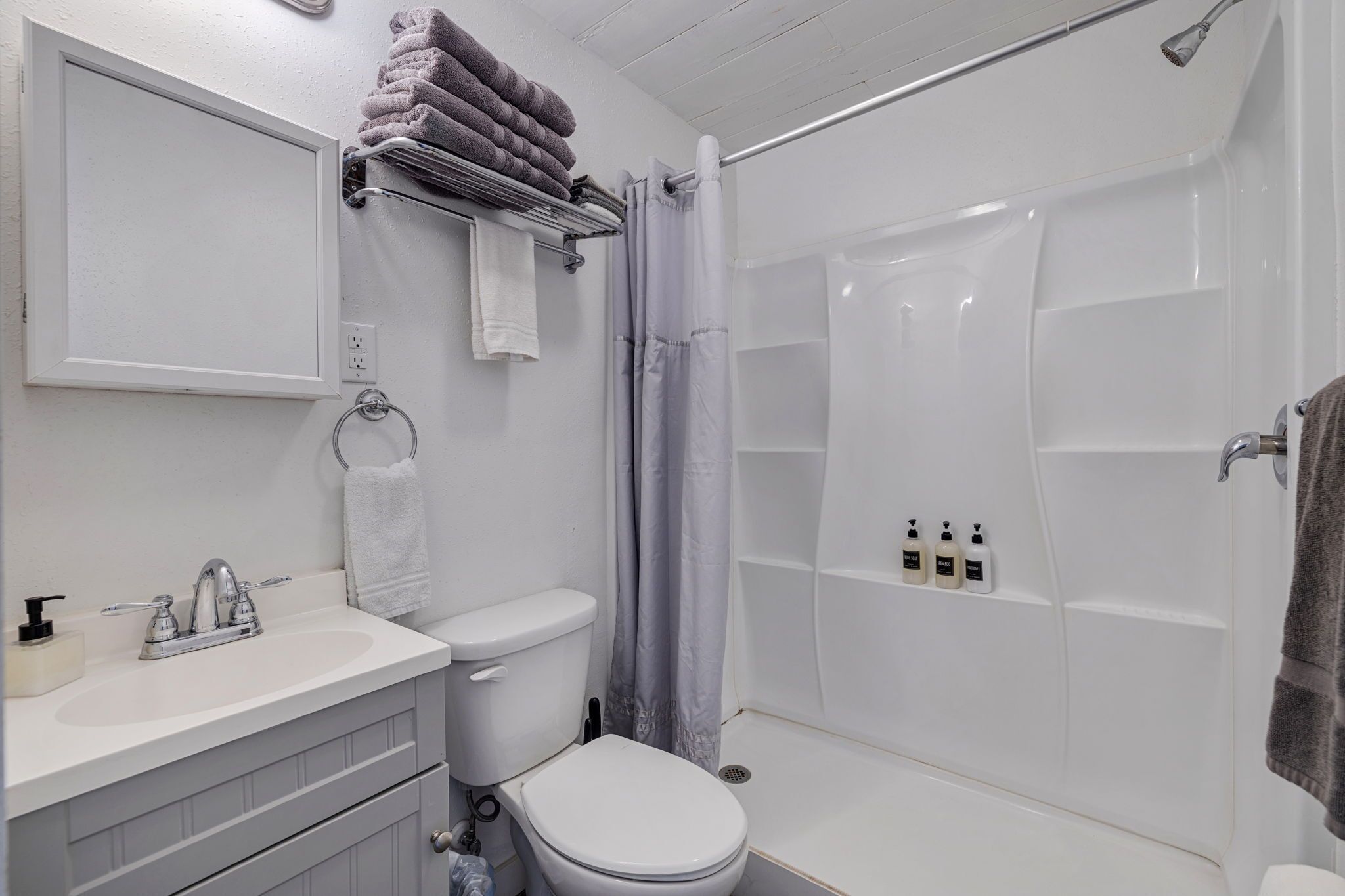 Un baño con lavamanos, inodoro y ducha blancos. Hay toallas en un perchero y artículos de aseo en la pared.