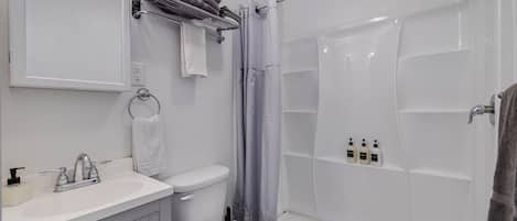 Un baño con lavamanos, inodoro y ducha blancos. Hay toallas en un perchero y artículos de aseo en la pared.
