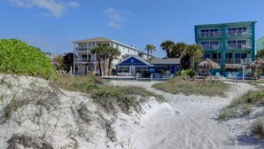 Una propiedad frente a la playa con un edificio de dos pisos, un restaurante en la playa y un área de piscina con sombrillas.