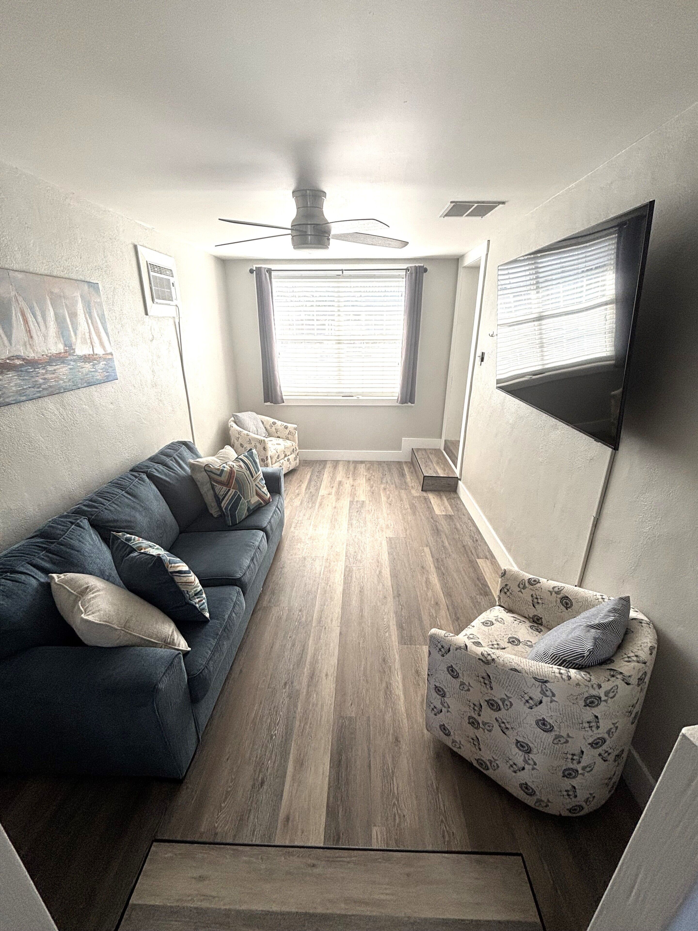 A compact living room with a blue sofa, patterned armchair, and a ceiling fan.