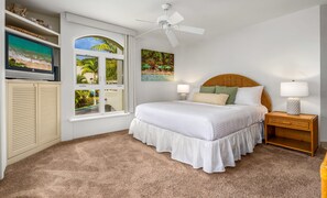 Une chambre à coucher comprenant un lit, deux tables de chevet, un ventilateur de plafond, une télévision et une fenêtre avec des volets.