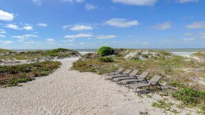 Um caminho de areia leva a cadeiras de praia, com vista clara do oceano e céu parcialmente nublado.