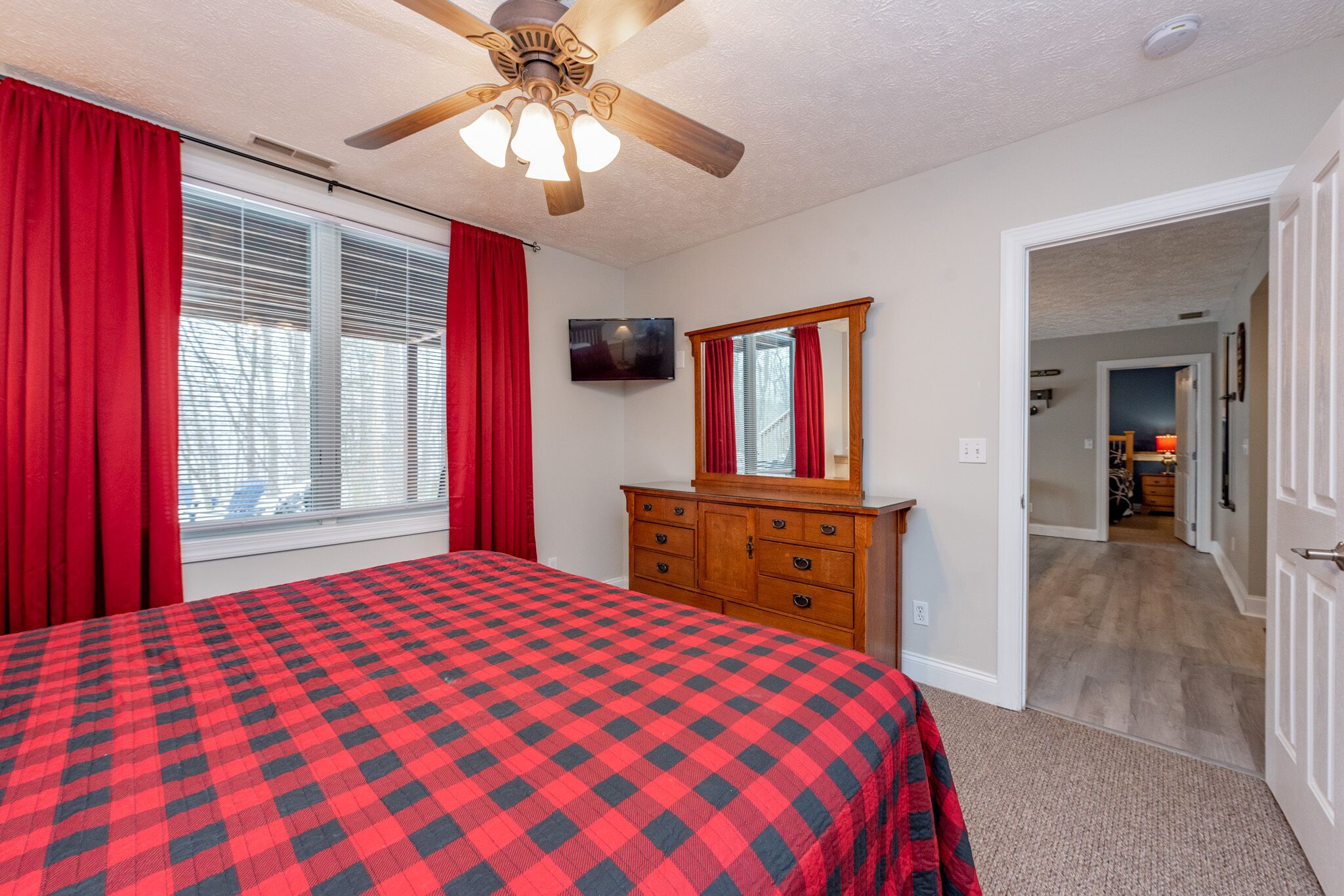 A bedroom with a checkered bedspread, a wooden dresser, a ceiling fan, and a television on the wall.