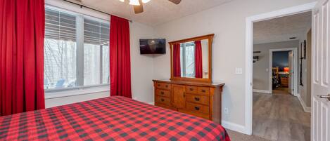A bedroom with a checkered bedspread, a wooden dresser, a ceiling fan, and a television on the wall.