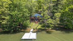 A house surrounded by dense forest, a dock extending into a body of water, and a staircase leading to the main entrance.