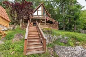 A wooden cabin with a staircase leading to a deck, surrounded by trees and rocks. - Rustic Chalet w/ Fireplaces & Jetted Tub (McHenry)