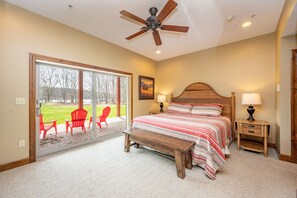 A bedroom with a bed, a wooden headboard, two bedside tables with lamps, a ceiling fan, and a sliding glass door leading to an outdoor patio with red chairs.