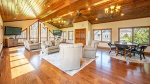 A spacious living room with a high ceiling, wooden beams, and large windows offering a view of the outdoors. - Luxury Lake Escape w/ Indoor Pool & Hot Tubs (Swanton)