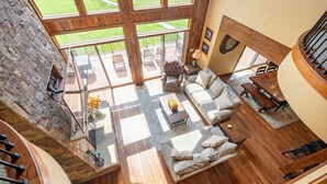 A spacious living area with a large window, a fireplace, and a wooden staircase. - Luxury Lake Escape w/ Indoor Pool & Hot Tubs (Swanton)