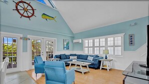 A living room with a blue sofa, matching armchairs, a coffee table, and a TV stand. There is a large window with shutters, a door leading outside, and a ceiling with a ship's wheel and fish decoration. - P78 - Charming 3 bedroom 2 bath home in Sombrero Beach with dock and pool (Marathon)