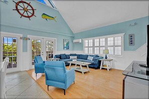 A living room with a blue sofa, matching armchairs, a coffee table, and a TV stand. There is a large window with shutters, a door leading outside, and a ceiling with a ship's wheel and fish decoration.