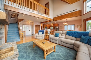 A spacious living area with a high ceiling, a staircase, and a kitchen in the background.