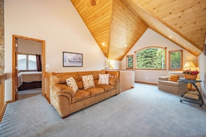 A loft-style room with a high wooden ceiling, a brown leather sofa, a grey armchair, and a small table with a lamp.