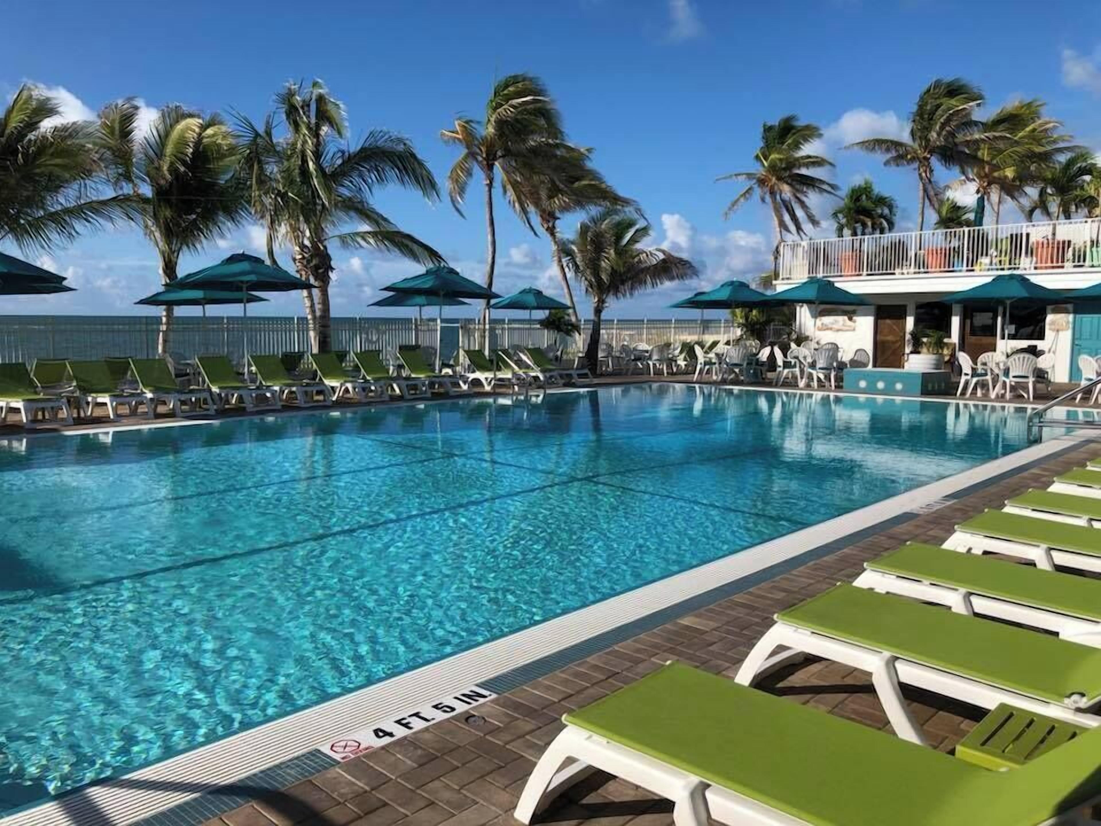 A swimming pool with clear blue water, surrounded by lounge chairs and umbrellas, with palm trees and a building in the background.
