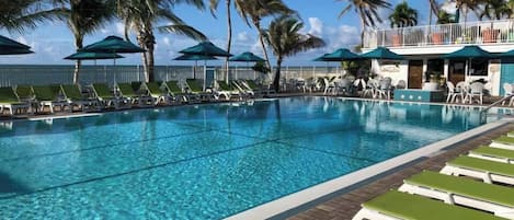 A swimming pool with clear blue water, surrounded by lounge chairs and umbrellas, with palm trees and a building in the background.