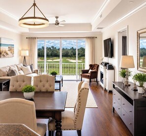 A spacious living room with a large window offering a view of a golf course, a dining area with a wooden table, and a seating arrangement with a sofa and chairs.