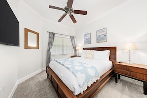 Quarto com cama de madeira, ventoinha no teto, janela com cortinas, abajur e duas fotos emolduradas na parede.