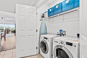 A laundry room with two front-loading washing machines, a drying machine, and blue storage bins labeled 'clean' and 'laundry.' - Beautiful Modern Condo with Private Balcony and Golf Course Views (Four Corners)
