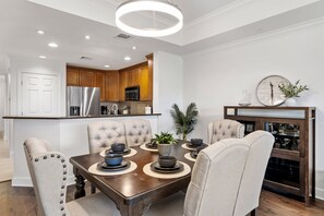 A modern dining area with a wooden table, upholstered chairs, and a kitchen in the background. - Beautiful Modern Condo with Private Balcony and Golf Course Views (Four Corners)