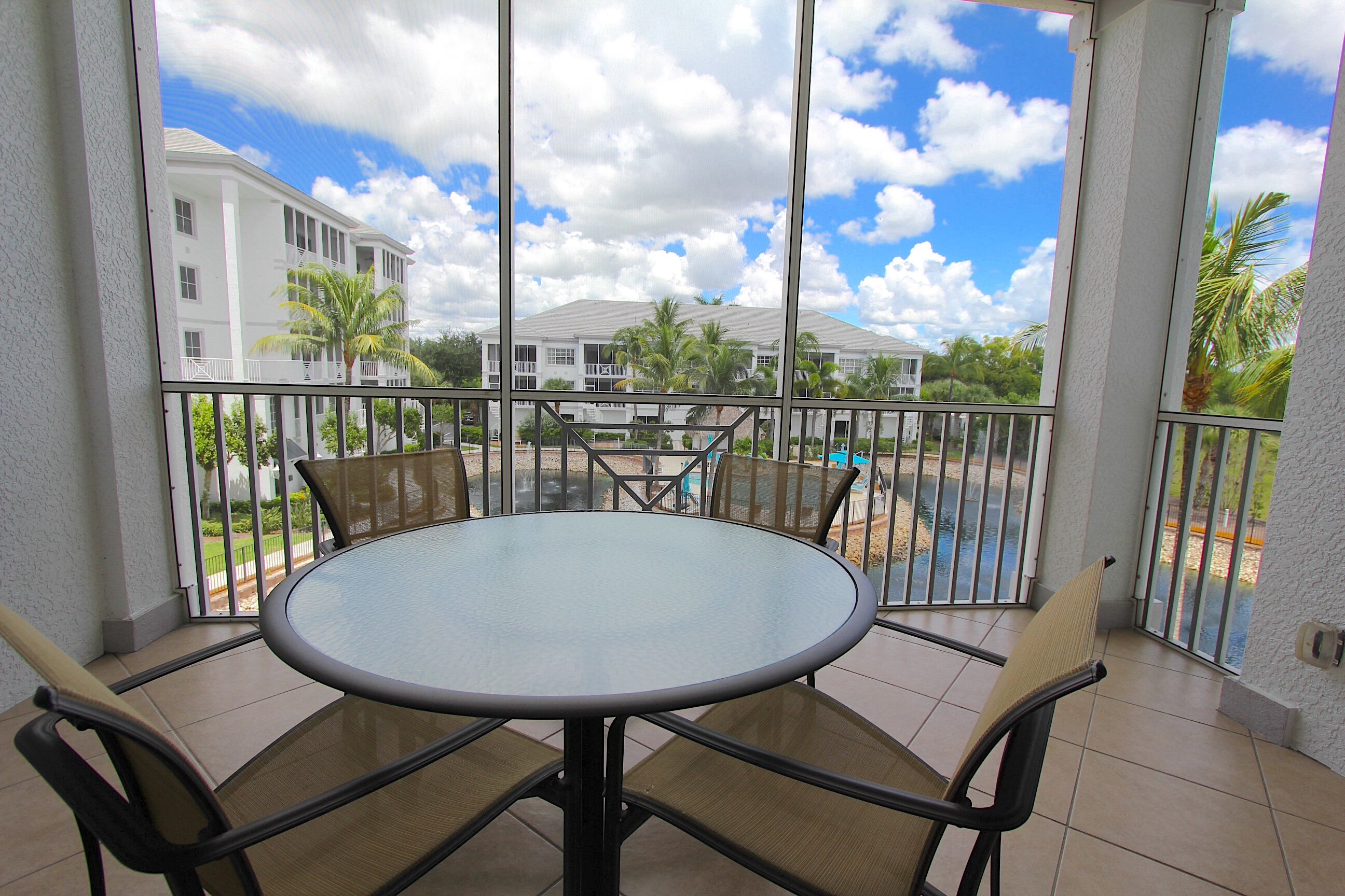 A balcony with a glass table and chairs, overlooking a swimming pool and a building with a white exterior.