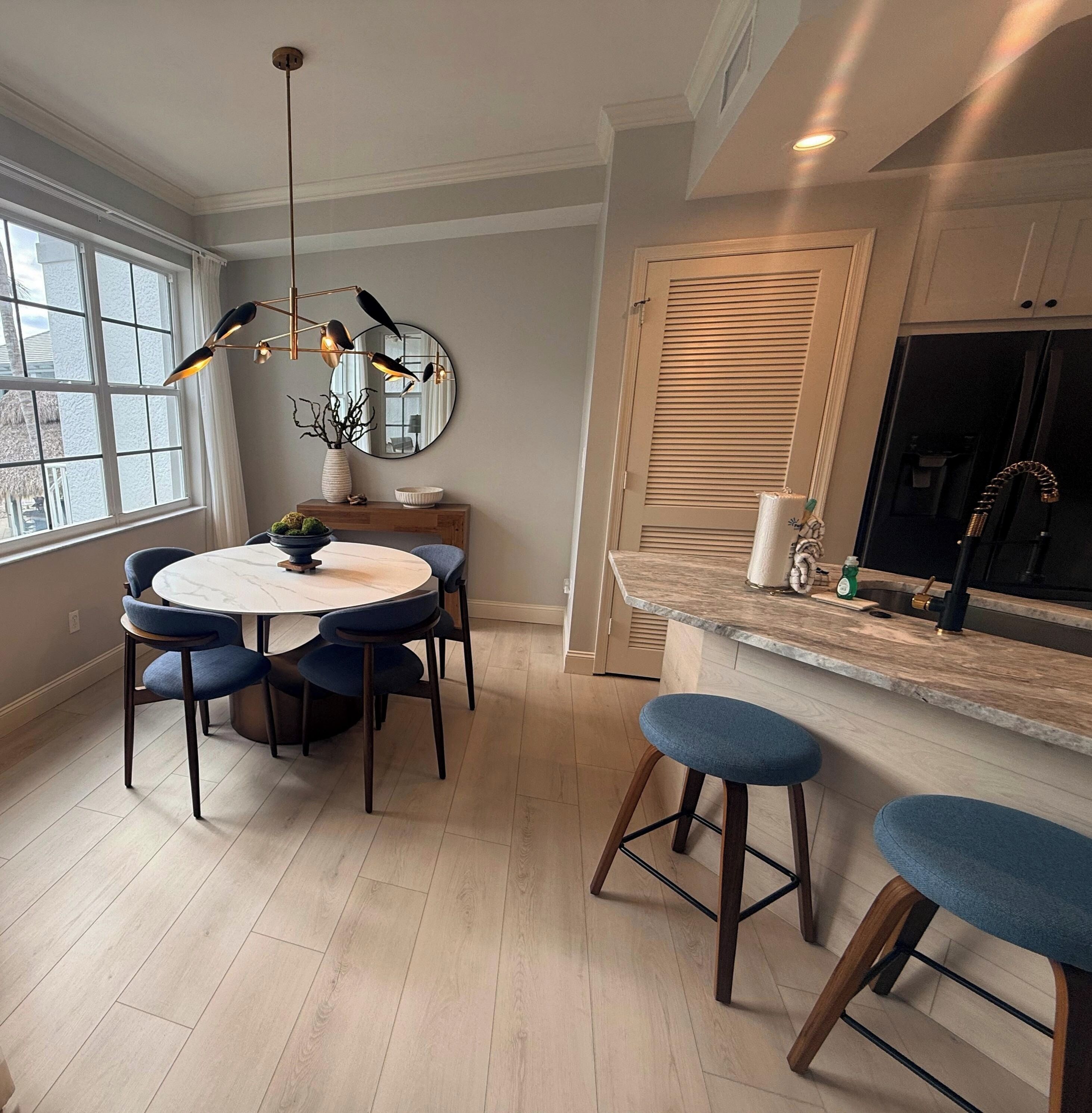 A modern kitchen with a round dining table, blue chairs, a hanging light fixture, and a large window.