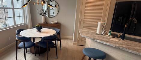 A modern kitchen with a round dining table, blue chairs, a hanging light fixture, and a large window.