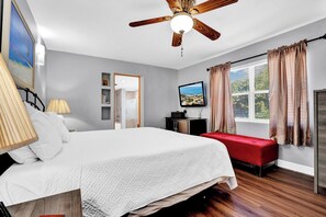 A bedroom with a bed, a red bench, a ceiling fan, a TV, and a window with curtains.