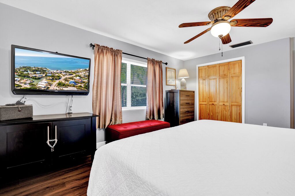 Ein Schlafzimmer mit einem Bett, einem Fernseher, einem Deckenventilator, einem Fenster mit Vorhängen und einem Kleiderschrank.