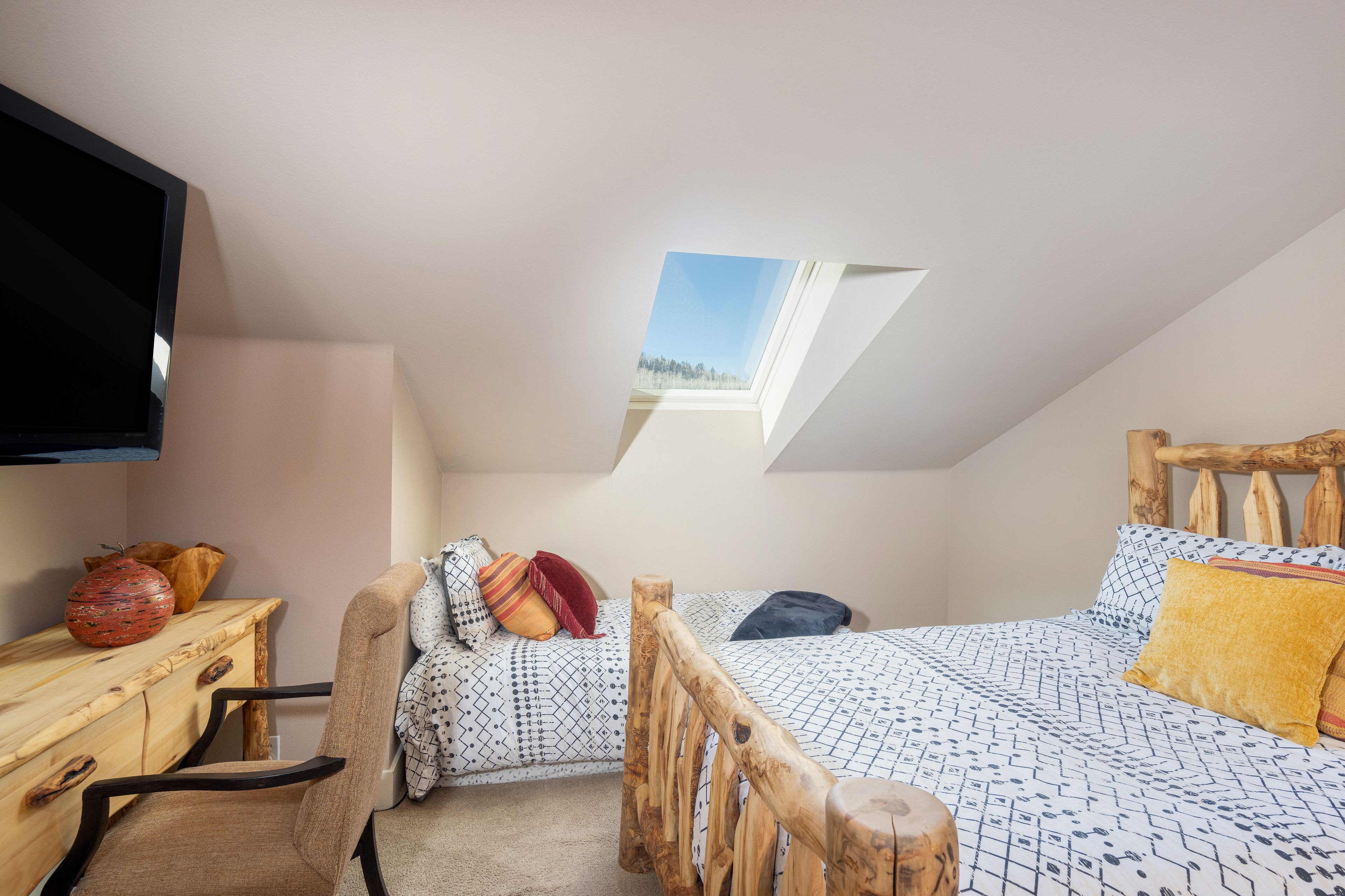 Un dormitorio en el ático con tragaluces, una cama con ropa de cama estampada, cabecero de madera, un escritorio con un jarrón y una silla.