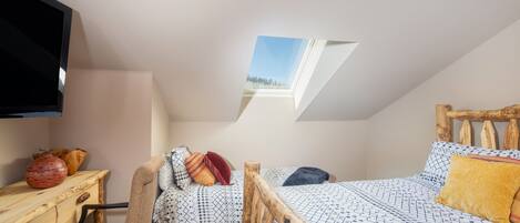 Un dormitorio en el ático con tragaluces, una cama con ropa de cama estampada, cabecero de madera, un escritorio con un jarrón y una silla.