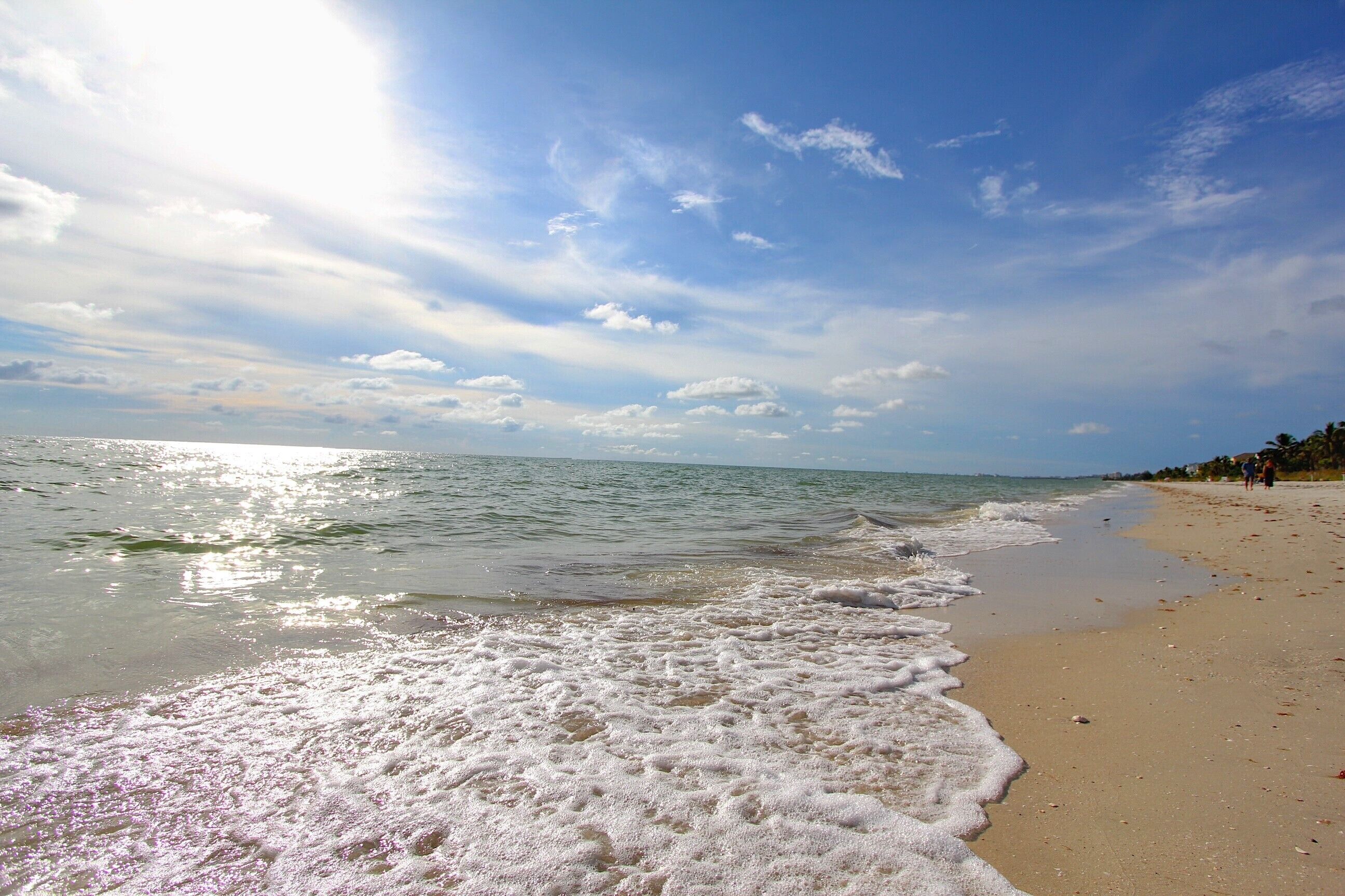 A beach with waves gently lapping onto the shore, a clear sky, and a few people in the distance.