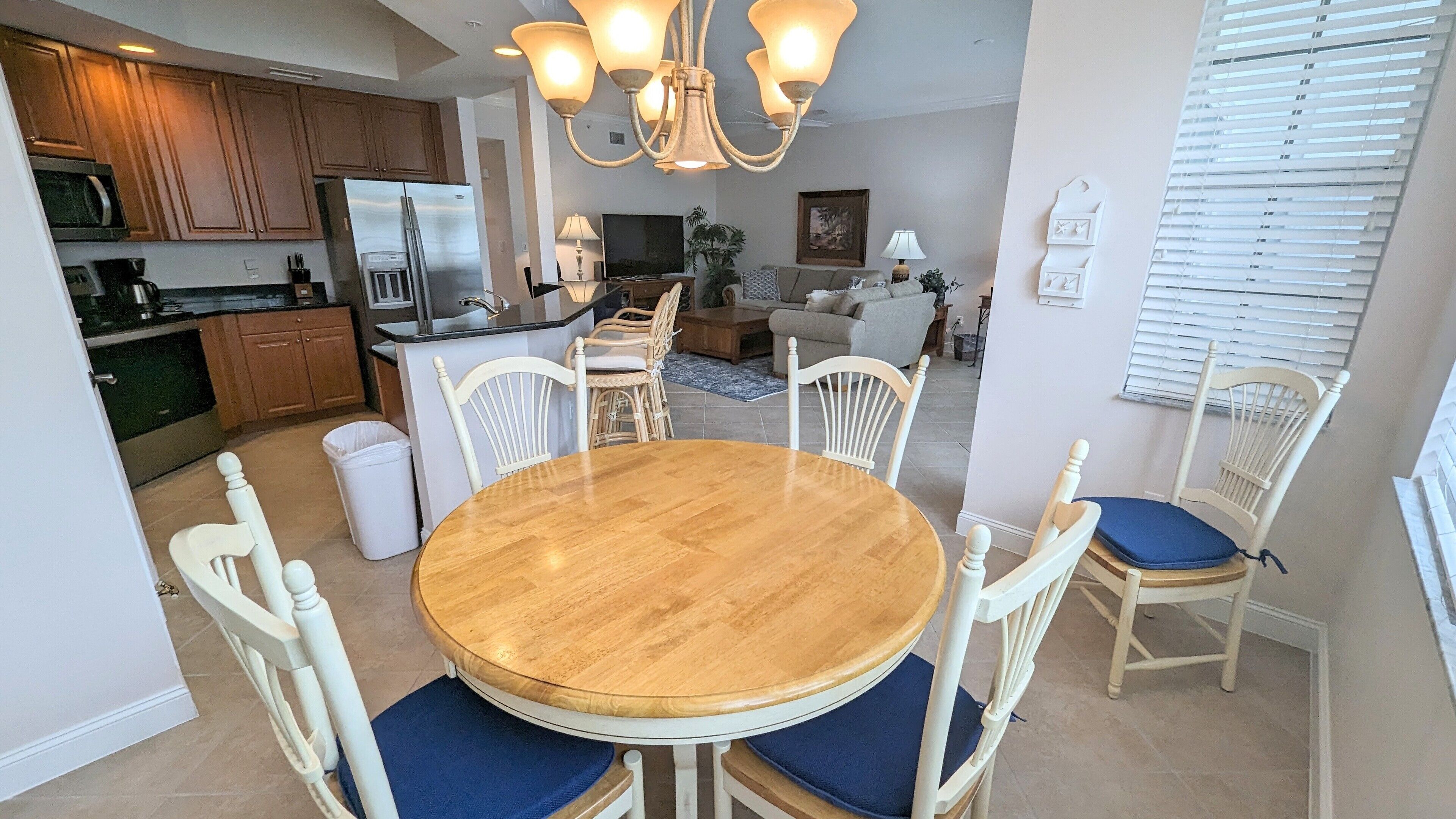 Un coin repas avec une table ronde en bois et des chaises blanches dotées de coussins bleus, une cuisine avec des meubles en bois et un réfrigérateur en acier inoxydable, et un salon avec un canapé et une table basse.