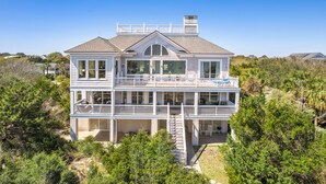 Une maison à deux étages située en bord de mer, dotée de plusieurs balcons, d’un porche couvert et d’un escalier menant à l’étage supérieur.