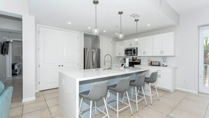 Una cocina moderna con armarios blancos, una isla central con taburetes de bar, electrodomésticos de acero inoxidable e iluminación colgante.