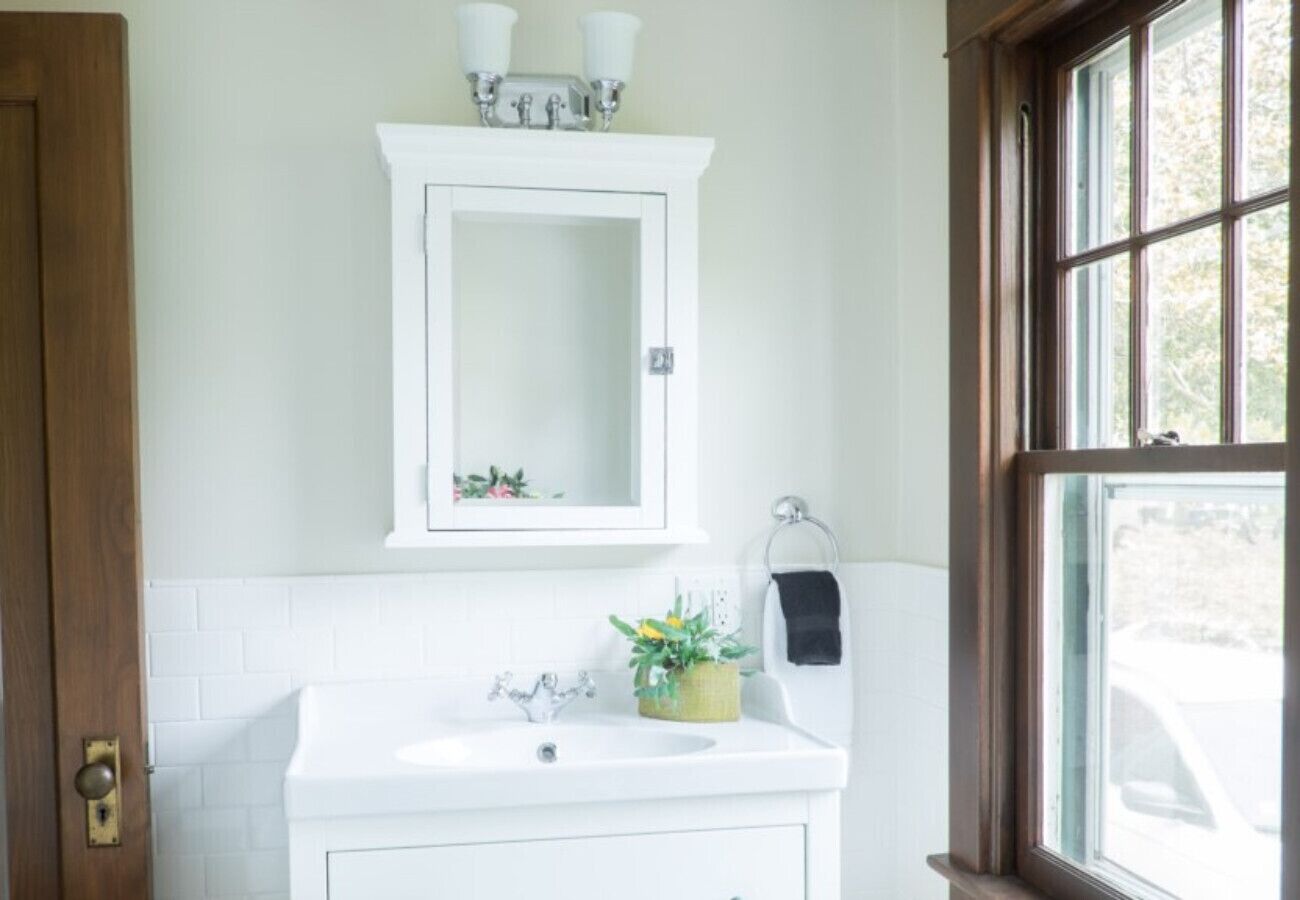 Un bagno con un lavandino bianco, uno specchio sopra e una finestra a destra.