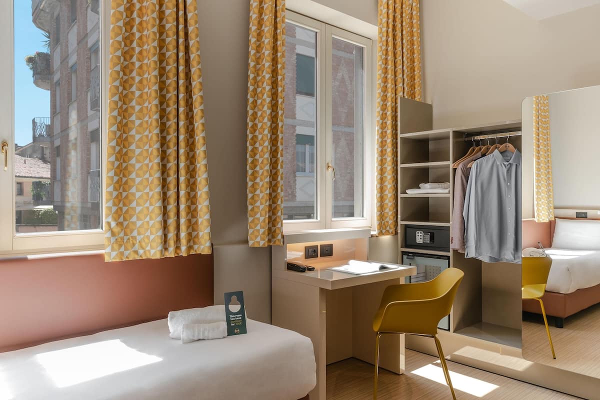 A hotel room with a bed, desk, chair, wardrobe, and a view of the city.