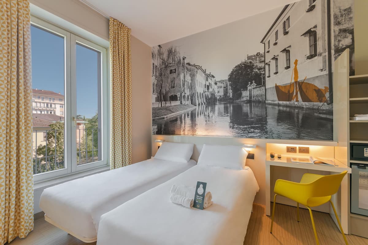 A hotel room with two beds, a desk, a chair, and a large mural of a canal scene.