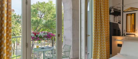 A hotel room with a bed, a small table with chairs, a balcony with a table and chairs, and a view of trees and flowers outside.