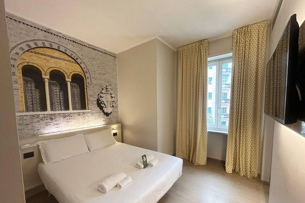 A hotel room with a large bed, a brick wall with a clock, and a window with curtains.