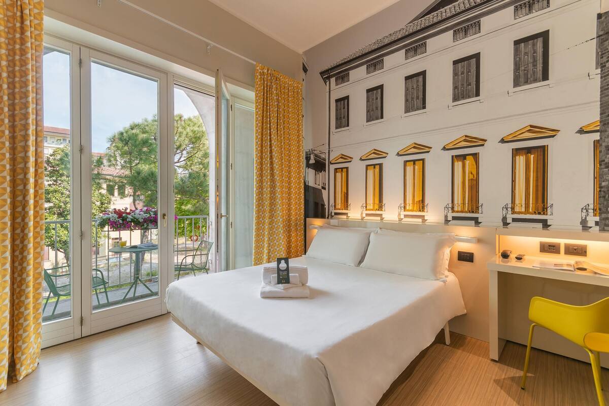 A hotel room with a bed, a desk, and a balcony with yellow curtains.