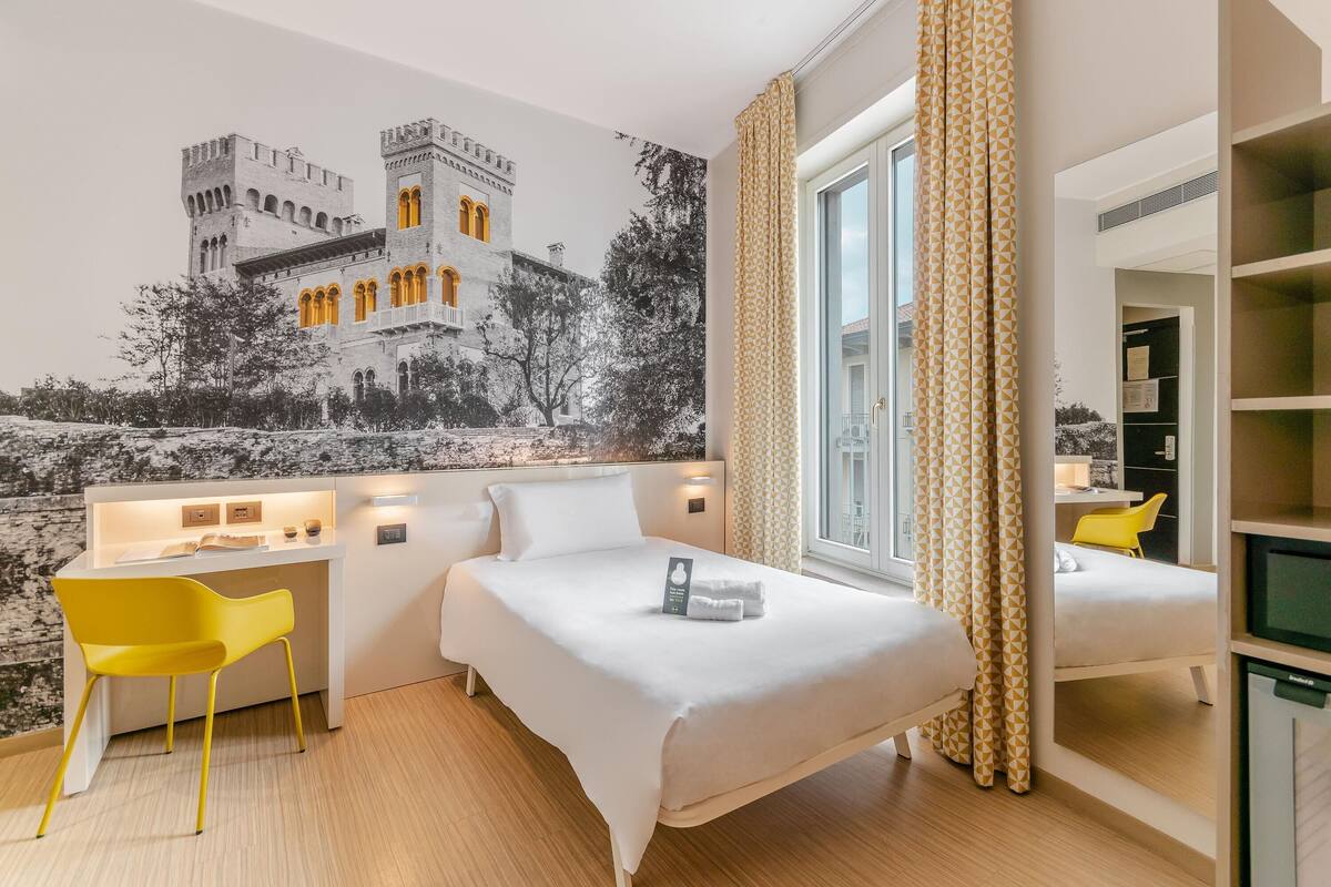 A hotel room with a bed, a desk, a chair, a large window, and a wall mural.