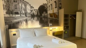 A hotel room with a bed, a nightstand, a mirror, a wardrobe, and a large wall mural of a canal scene. - B&B Hotel Treviso (Treviso)